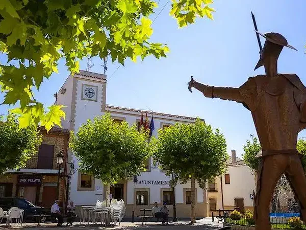 Escultura de Don Quijote frente al Ayuntamiento de Ossa de Montiel, en una plaza soleada con terrazas y árboles, destino turístico de la Ruta del Quijote.