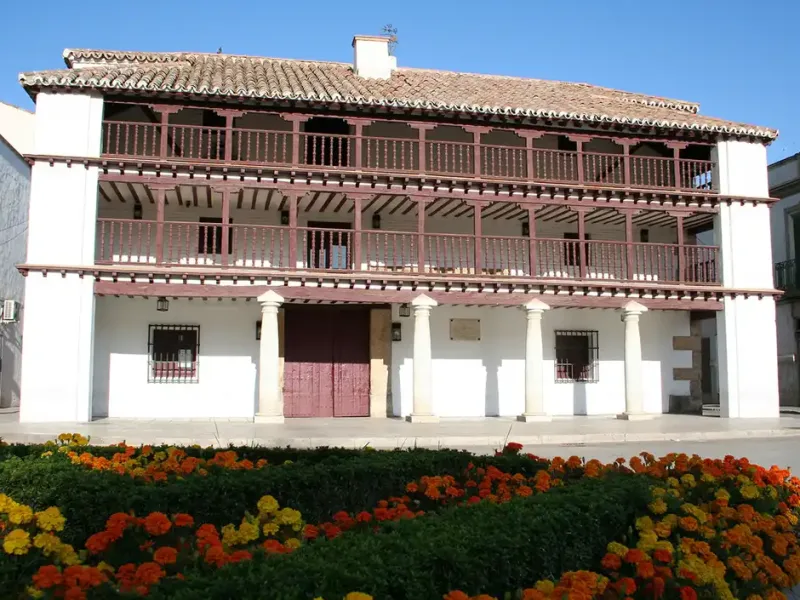 Fachada del antiguo granero municipal de Tomelloso, actual teatro-auditorio, con estructura de madera y columnas, frente a un parterre de flores coloridas.